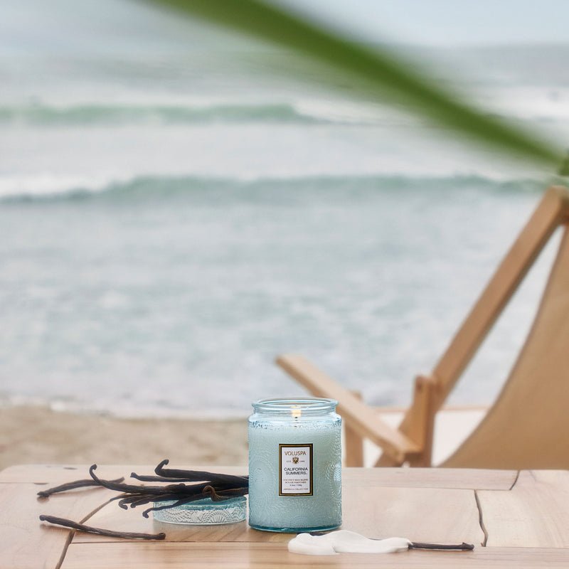 The Voluspa - California Summers Small Jar Candle 5.5oz casts a serene glow from its coconut wax blend on a wooden table. Dried vanilla pods are scattered nearby with a tranquil beach and deck chair in the background, while waves create refreshing aquatic notes under an overcast sky.