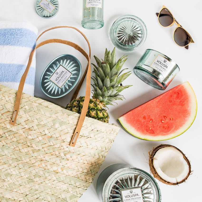 A summer-themed setup includes a straw bag, striped towel, pineapple, watermelon slice, half coconut, and sunglasses. Several glass candles and a Voluspa - Casa Pacifica 192ml Reed Diffuser enhance the beach or picnic atmosphere with an inviting fragrance.