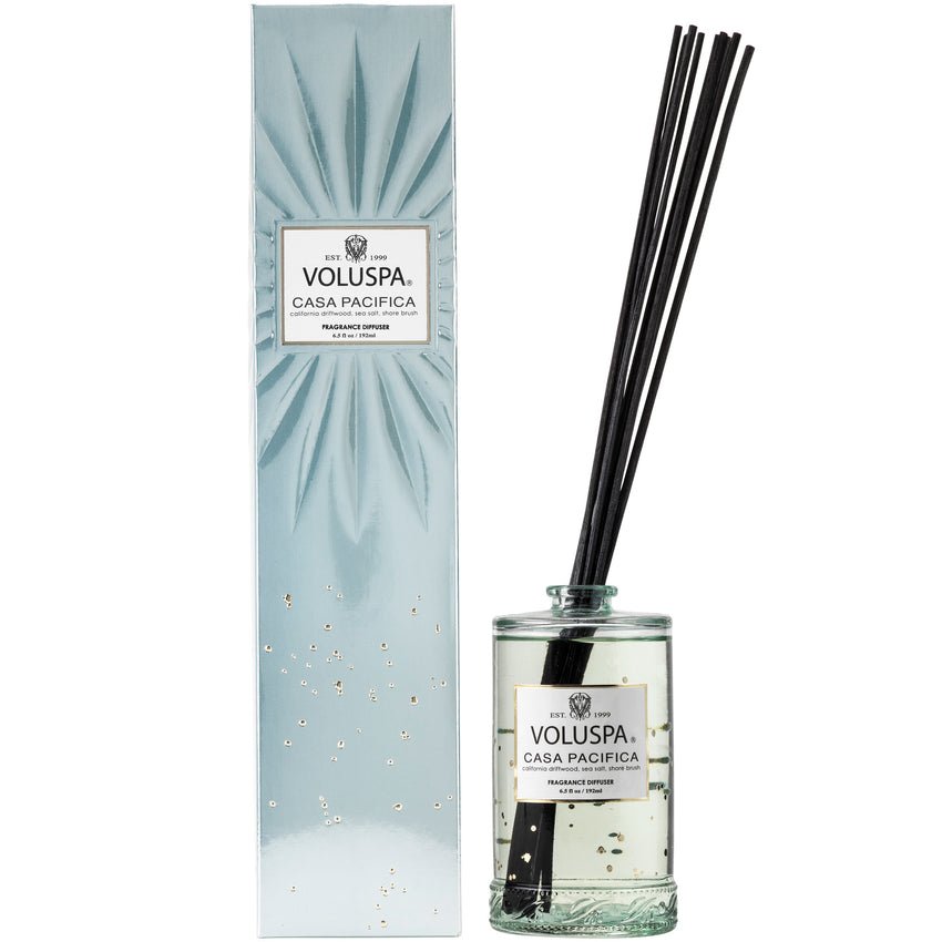 The Voluspa Casa Pacifica 192ml Reed Diffuser in a clear glass jar with black sticks elegantly disperses fragrance oil. The matching boxed set features a blue-green design with gold speckles, making it a chic room accent.