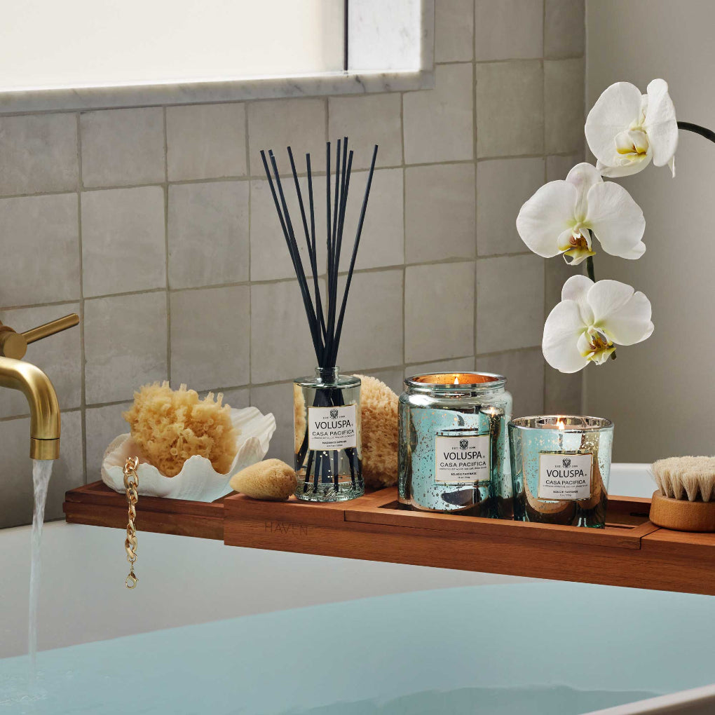 A bathtub scene includes a wooden tray with a Voluspa - Casa Pacifica Large Jar 18oz Candle, a reed diffuser, loofah, sponge, and white orchid. The tub is filled under a gold faucet against a tiled wall.