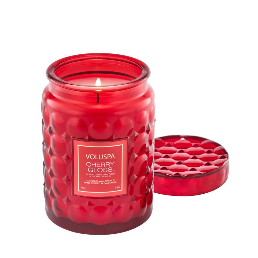 The Voluspa Cherry Gloss Large Jar 18oz Candle is lit, emitting a warm glow through its textured glass. The lid with matching design sits nearby. Made from luxurious coconut wax, this candle fills the room with an inviting aroma.