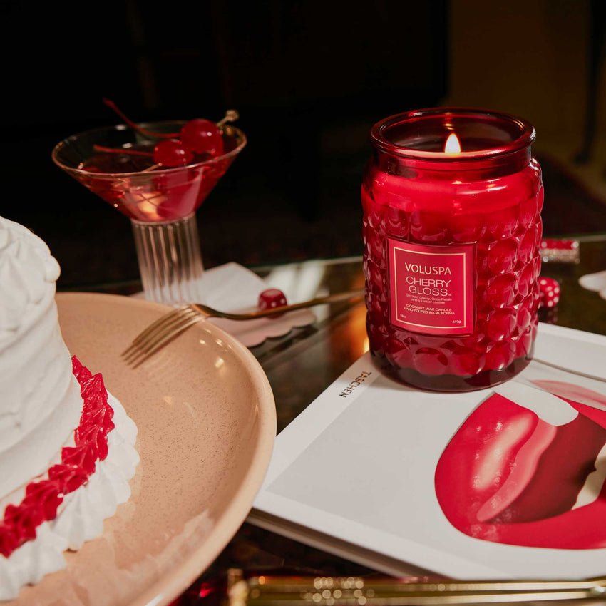 A Voluspa Cherry Gloss Large Jar 18oz Candle sits on a table next to a slice of white cake with red frosting, an open magazine, and a cocktail glass with cherries. Made from coconut wax blend, its perfect for creating a cozy, intimate atmosphere.