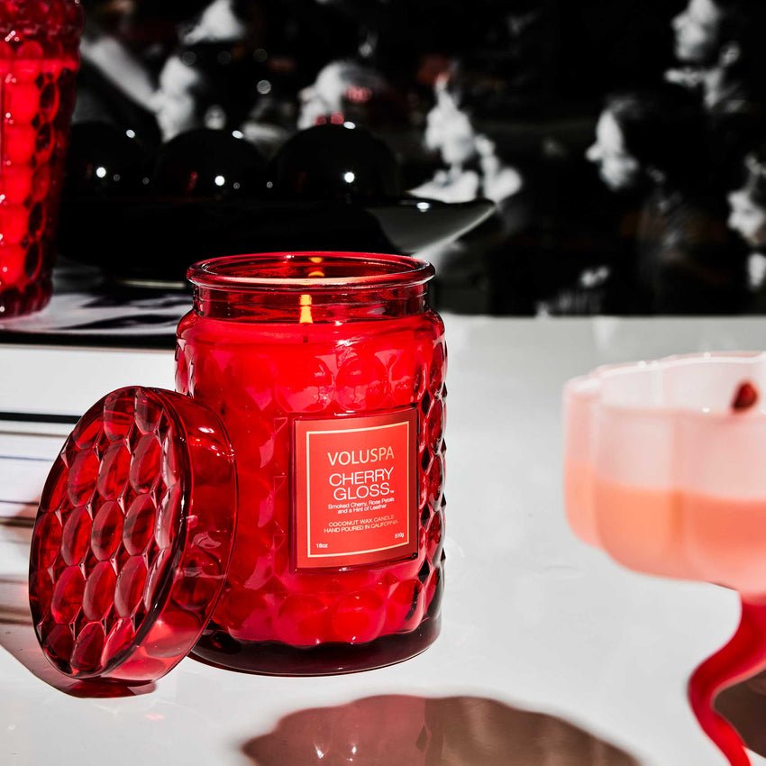 The Voluspa - Cherry Gloss Large Jar 18oz Candle, crafted with a coconut wax blend in a textured glass jar, illuminates the table while dark blurred elements in the background contrast with its radiant glow.