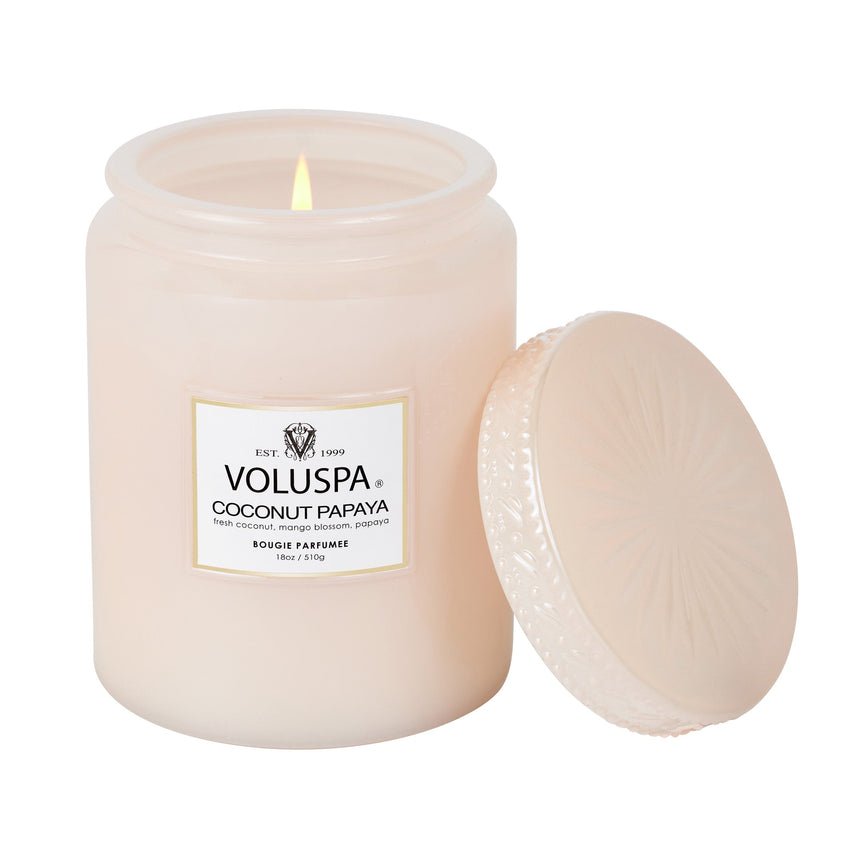 The lit Voluspa Coconut Papaya Large Jar 18oz Candle, with its lid leaning against the frosted glass jar, releases a soothing tropical fruit fragrance. The label shows the scent and brand logo, inviting you to enjoy its coconut wax blends aroma.