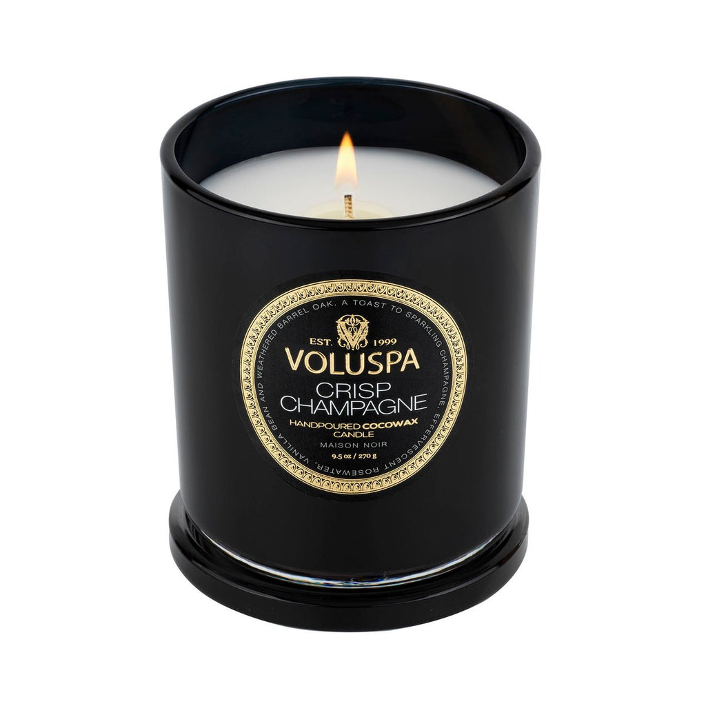 The 9.5oz Voluspa Crisp Champagne candle, made from a coconut wax blend, is housed in a sleek black glass jar with a gold and black label. It sits on its matching lid, showcasing the creamy white hue of the hand-poured candle.