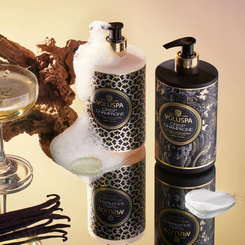 Two elegant Voluspa Crisp Champagne soap dispensers and a Voluspa Crisp Champagne Hand Lotion sit on a reflective surface with soap bubbles. A glass of champagne, vanilla pods, wood, and a white petal-like object complete the scene, creating a luxurious setting.