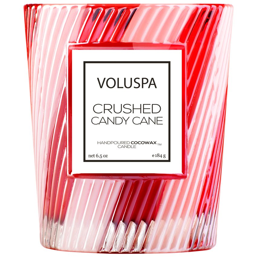 The Voluspa - Crushed Candy Cane Classic 6.5oz Candle features a pink and red striped glass jar with a white label, and contains hand-poured coconut wax in the Crushed Candy Cane scent.