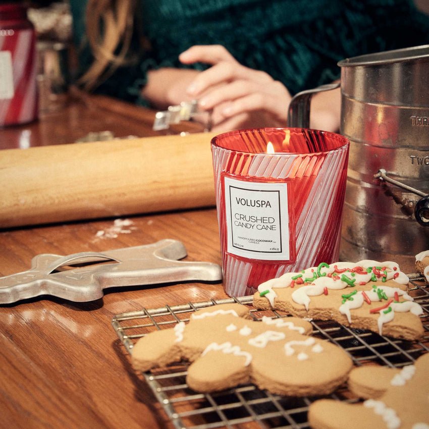 A lit Voluspa Crushed Candy Cane Classic 6.5oz Candle sits on a wooden table beside gingerbread cookies, a rolling pin, and a sifter, while hands in the background suggest festive holiday baking.