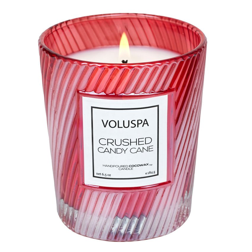 The Voluspa Crushed Candy Cane Classic 6.5oz Candle glows in a red, ribbed glass jar, filling the air with festive fragrance as its white coconut wax melts and the flame flickers softly at the center.