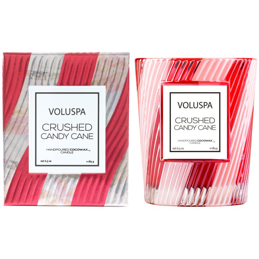 A Voluspa Crushed Candy Cane Classic 6.5oz Candle in pink and white stripes sits beside its matching box, both adorned with festive swirled patterns and labeled with the Voluspa brand.