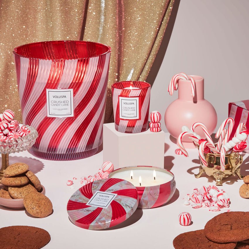 A Voluspa Crushed Candy Cane Classic 6.5oz Candle sits open on a festive table with cookies, candy canes, peppermints, pink decor, and a gold glittery backdrop.