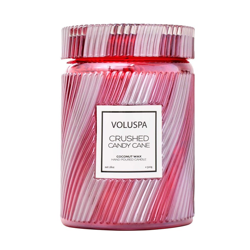 A pink and white ribbed glass jar holds the Voluspa - Crushed Candy Cane Large Jar 18oz Candle, a hand-poured coconut wax candle with a delightful crushed candy cane scent.