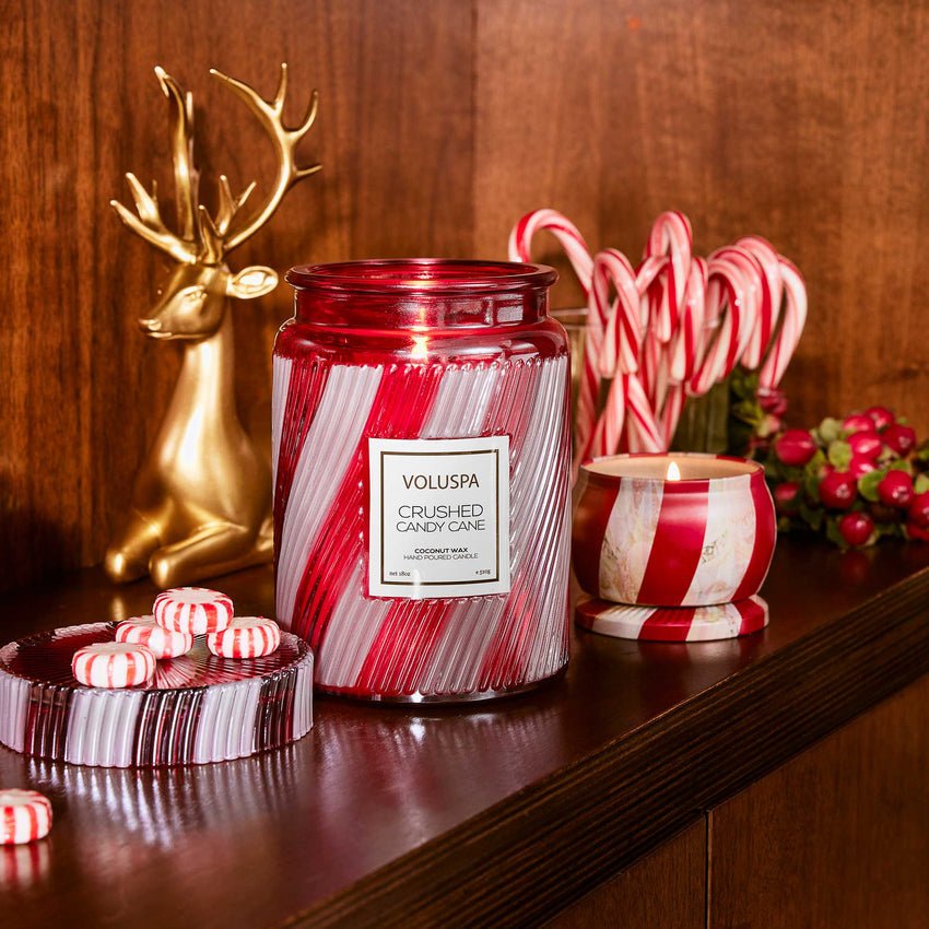 A festive shelf display features the Voluspa Crushed Candy Cane Large Jar 18oz Candle, peppermint candies, a red and white striped cup, candy canes, red berries, and a gold reindeer figurine set against a wooden background.