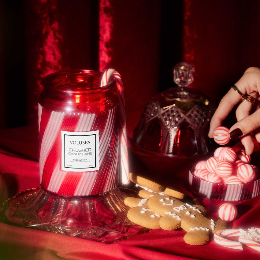A Voluspa - Crushed Candy Cane Large Jar 18oz Candle sits on a glass stand with striped candy canes, gingerbread cookies, and red-and-white candies over red velvet as a hand reaches for a treat.