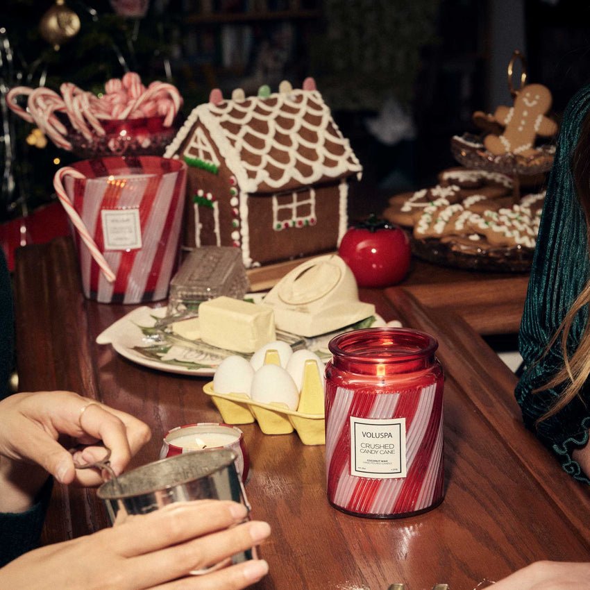 A cozy holiday scene features a Voluspa - Crushed Candy Cane Large Jar 18oz Candle, gingerbread house, candy canes, eggs, butter, and cookies on a wooden table as two people prepare festive treats together.