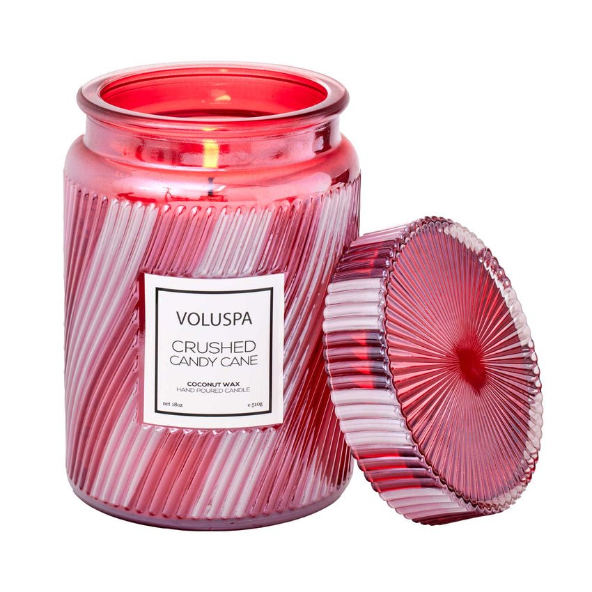 A pink and white ribbed glass jar labeled Voluspa - Crushed Candy Cane Large Jar 18oz Candle holds a lit peppermint candle made with coconut wax, with its matching lid open beside it. Brand: Voluspa.