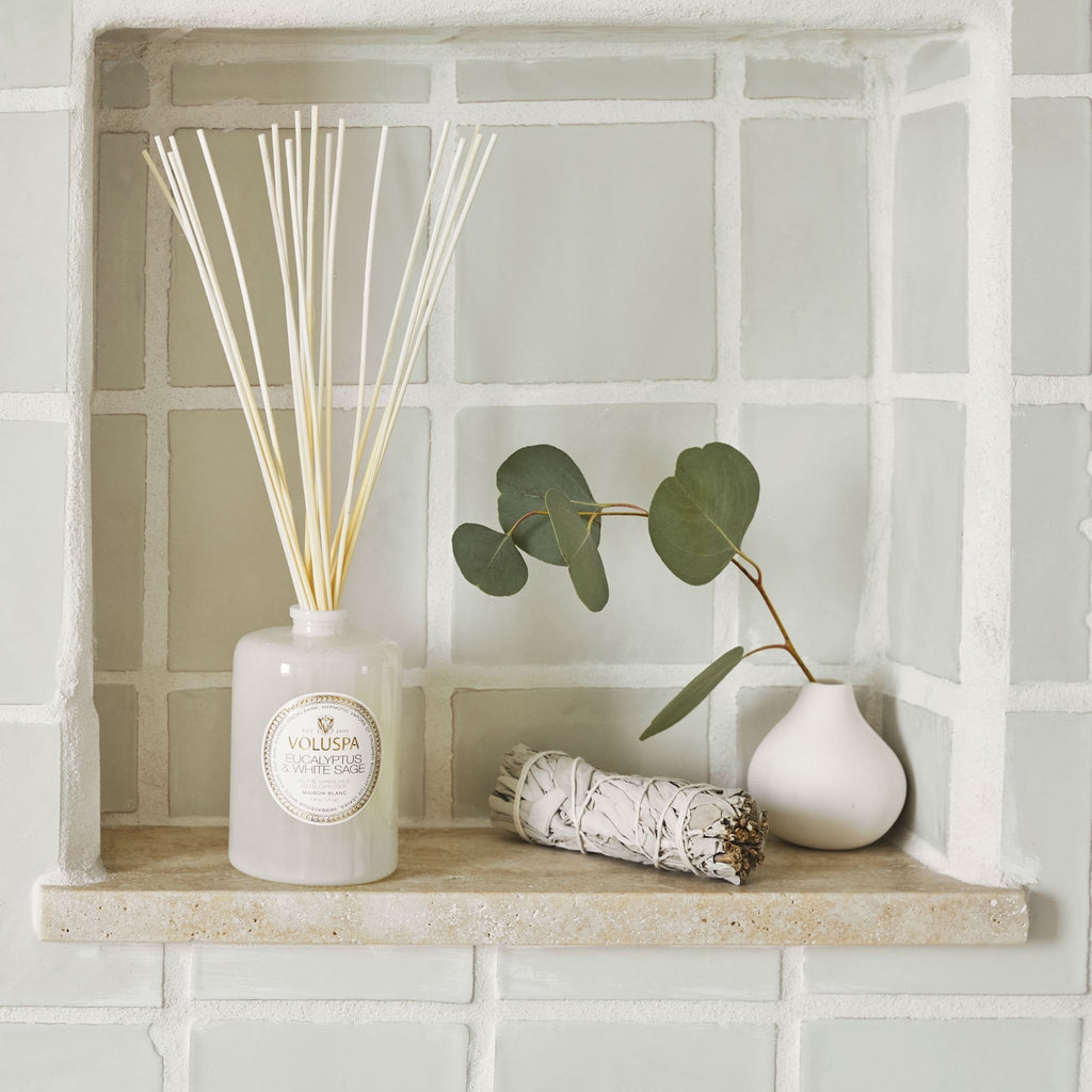 The Voluspa - Eucalyptus & White Sage 172ml Reed Diffuser with thin sticks sits elegantly on a tiled shelf beside a small white vase of Eucalyptus leaves, with a White Sage bundle set against textured light green and white tiles.