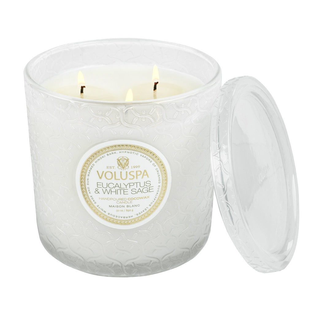 The Voluspa Eucalyptus & White Sage Luxe 30oz Candle showcases a textured glass design with a matching lid beside it. It features three wicks and emits calming eucalyptus and white sage scents.