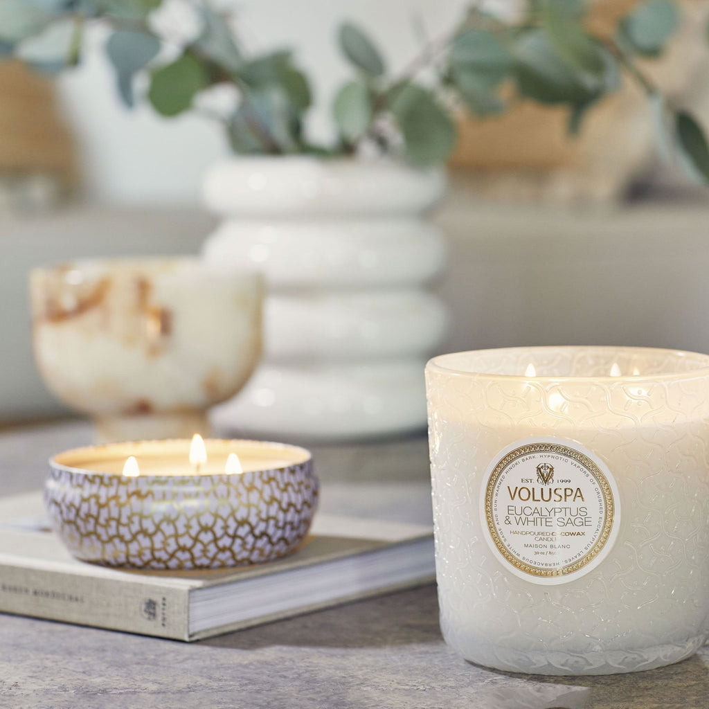 Two lit candles on the table, including a Voluspa Eucalyptus & White Sage Luxe 30oz candle in a textured white holder, create a cozy ambiance. Its soothing scent complements the decorative background candle beside a book and vase with greenery.