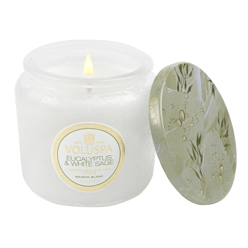 The Voluspa Eucalyptus & White Sage Petite 4.5oz Jar Candle emits soothing scents from its intricately embossed jar, topped with an ornate lid featuring green leaves and water droplets, elegantly placed by the gently flickering flame.