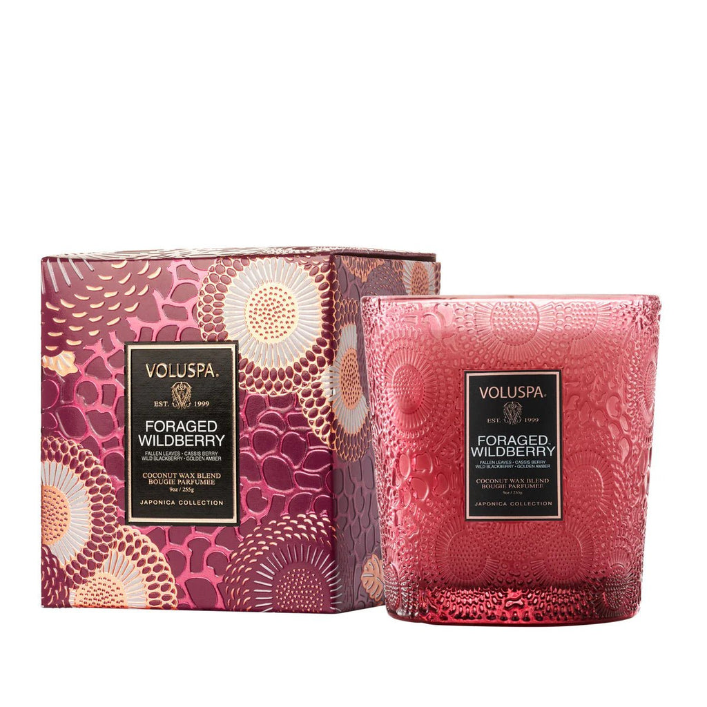 A decorative pink glass candle, Voluspa - Foraged Wildberry Classic Candle 9oz, features a citrus fragrance. It sits beside a matching patterned box with pink, gold, and burgundy floral design from the Japonica Collection. Made from coconut wax, its a vegan candle choice.