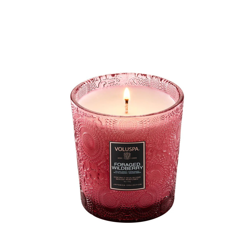 The Voluspa - Foraged Wildberry Classic Candle 9oz emits a hint of citrus from its textured pink glass holder. This coconut wax, vegan candle creates a warm glow against the white background as it gently melts.