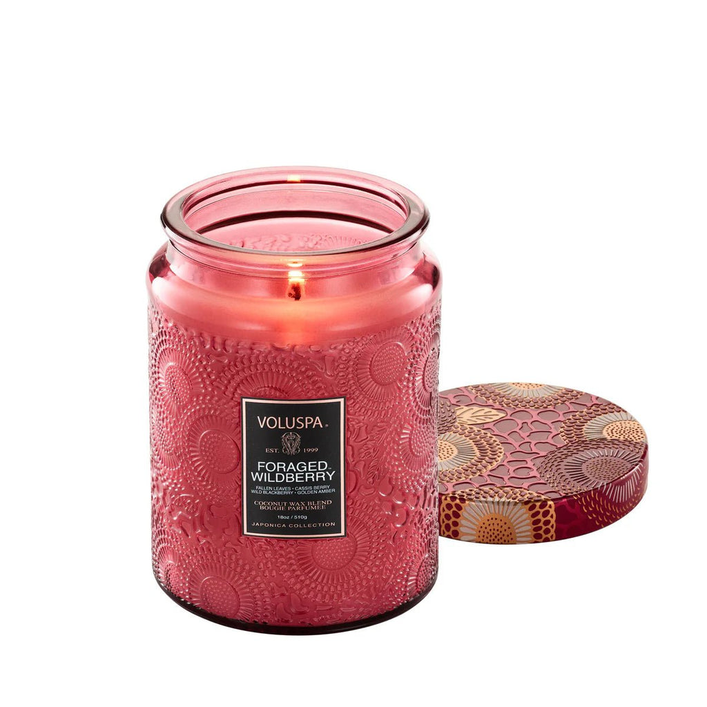 A lit Voluspa Foraged Wildberry 18oz candle in pink glass with a gold and red floral lid fills the room with a delightful citrus/fruity scent.