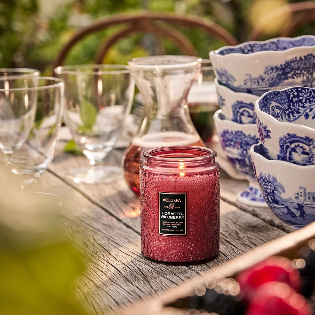 A Voluspa - Foraged Wildberry Small Jar 5.5oz Candle sits on a wooden outdoor table with glassware, a carafe, blue and white bowls, and fresh berries, its wildberry scent blending with the lush surroundings.