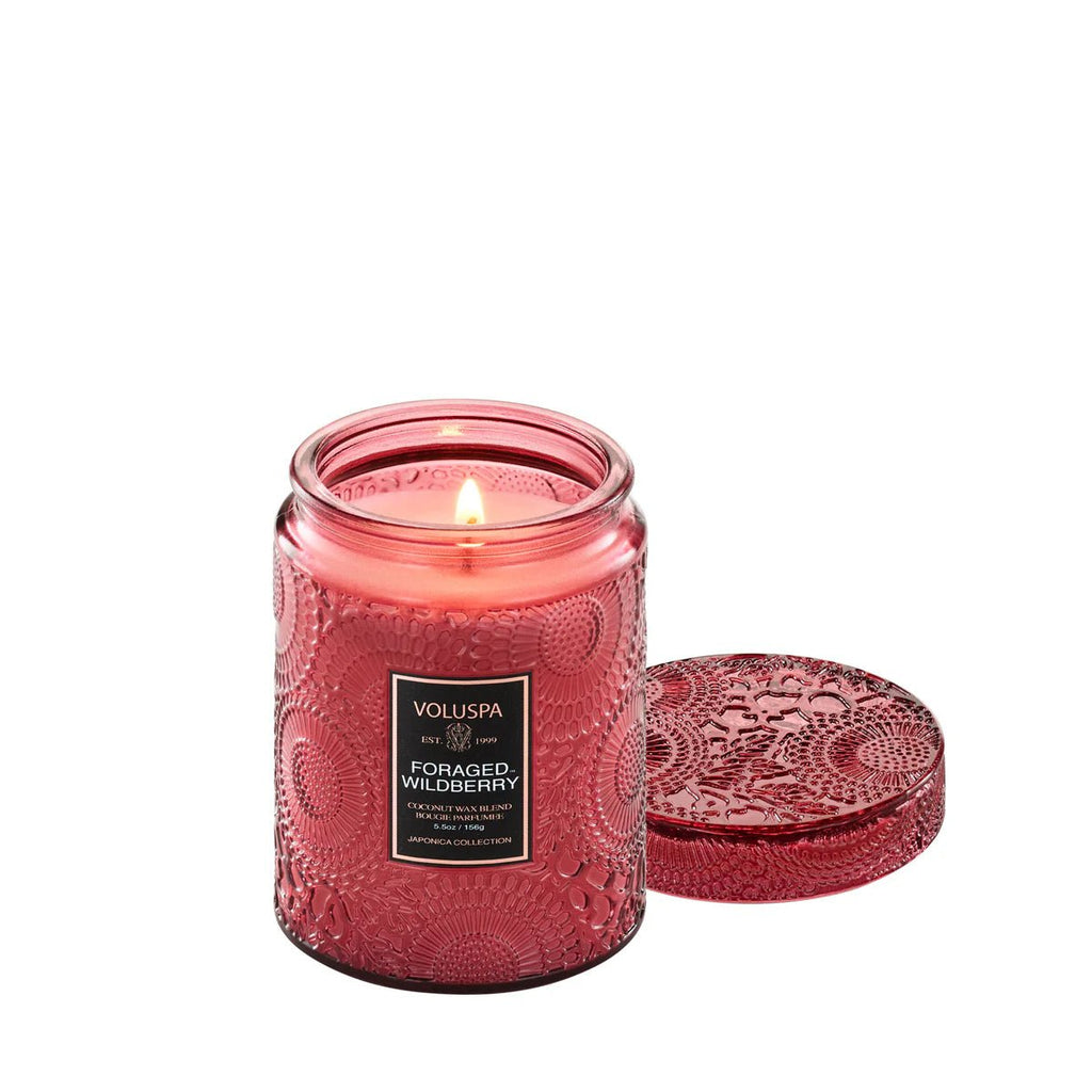 A lit Voluspa - Foraged Wildberry Small Jar 5.5oz Candle sits with its matching lid beside it, filling the air with a wildberry scent. The Voluspa label is clearly visible on the decorative pink jar.