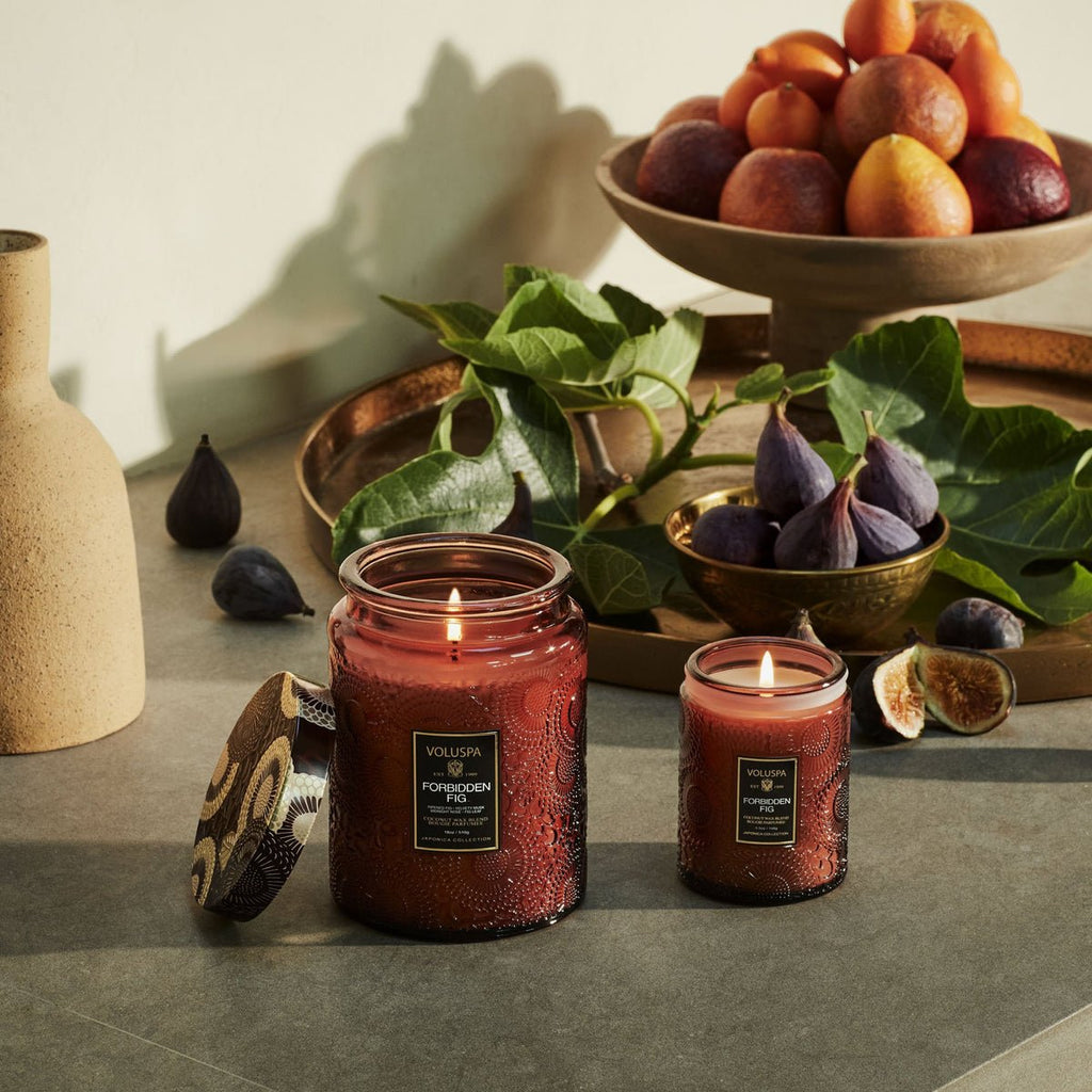 Two Voluspa Forbidden Fig Large Jar 18oz candles made from coconut wax flicker on a table with figs, tropical leaves, and a copper platter of ripe fruits. A beige vase adds rustic charm, completing the cozy and luxurious ambiance with velvety musk aromas in the air.