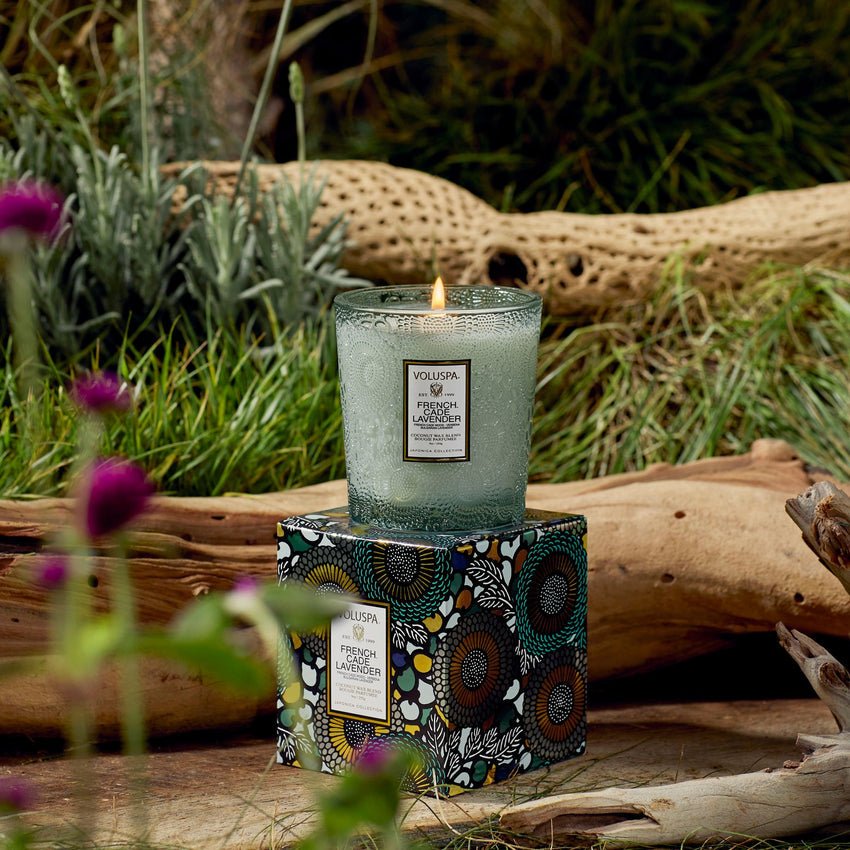 A lit Voluspa French Cade Lavender Classic 9oz Candle sits on its decorative box outdoors among greenery and flowers—a coconut wax blend candle with a relaxing lavender scent.
