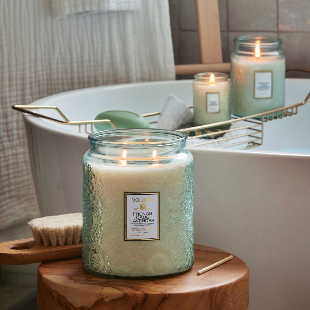 Voluspas French Cade Lavender Luxe Jar 44oz candle in glass jars, crafted from coconut wax, creates a relaxing bathtub atmosphere. A bath brush and matchstick rest on a wooden stool beside the tub.