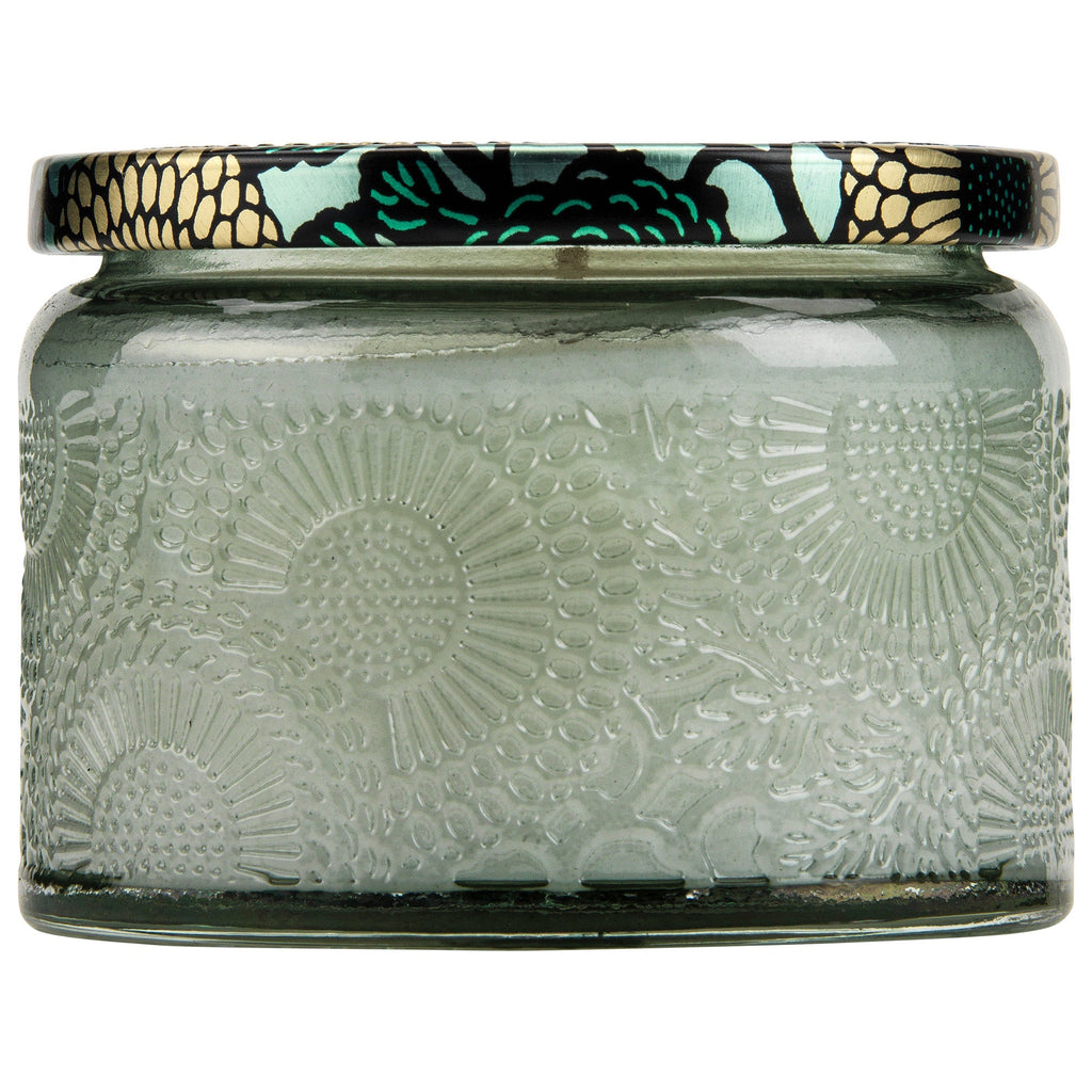 The Voluspa French Cade Lavender Petite 3.2oz Jar Candle features a decorative glass jar with an embossed floral pattern and a colorful lid showcasing a metallic floral design in shades of black, gold, and teal. It holds a blend of coconut wax embodying elegance.