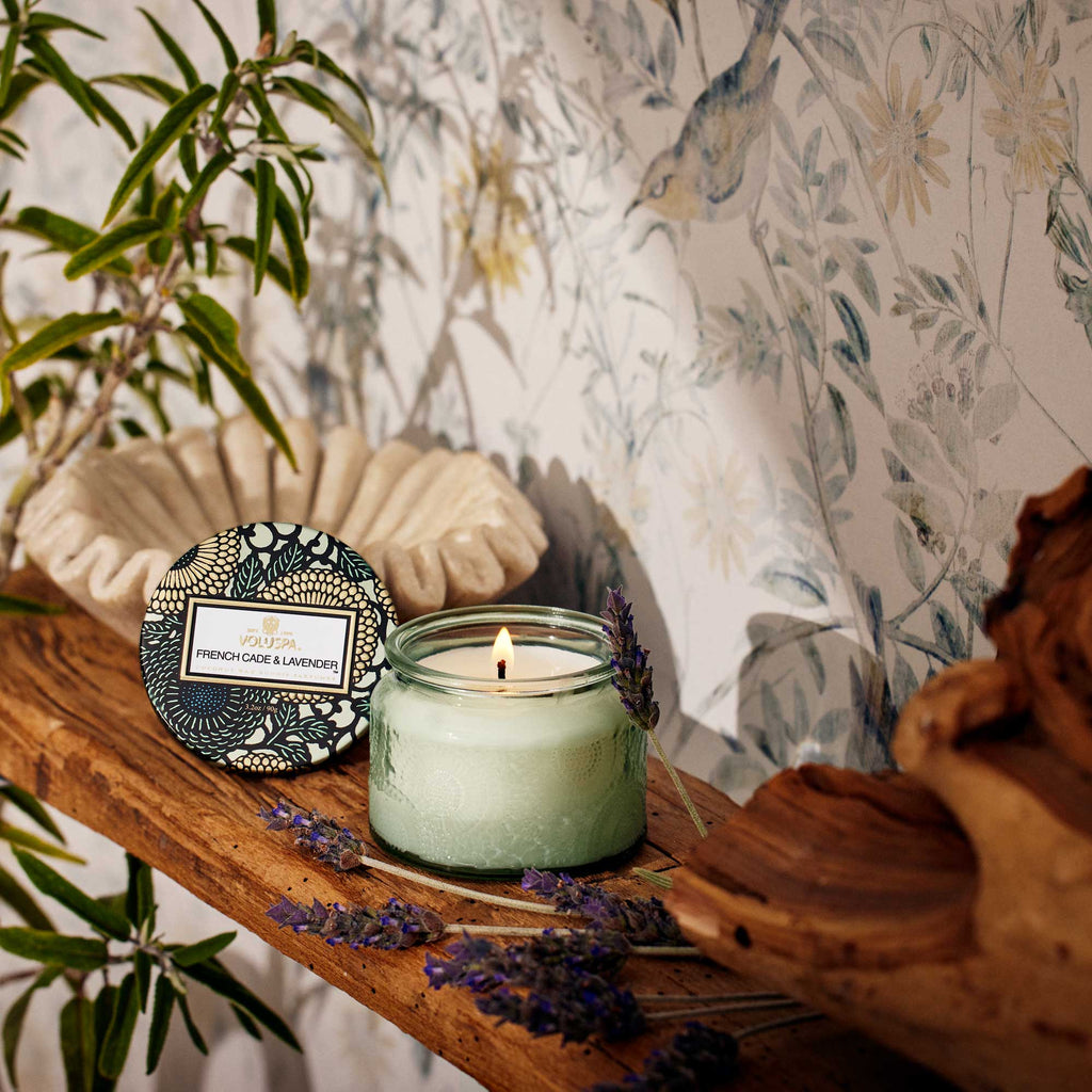 A lit Voluspa - French Cade Lavender Petite 3.2oz Jar Candle, made from a coconut wax blend, rests on a wooden shelf with a floral lid and lavender sprigs nearby. Floral wallpaper and a shell-shaped bowl set the backdrop, while plants partially frame this serene scene.