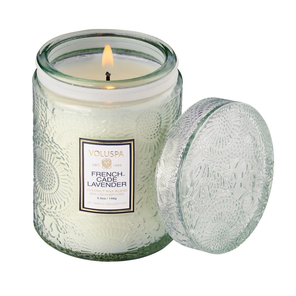 A lit Voluspa French Cade Lavender candle, 5.5oz with untouched pale coconut wax, sits in a floral-patterned jar. The lid beside it shares the embossed design.