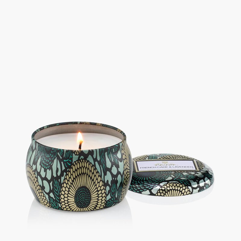 The Voluspa - French Cade Lavender Tin 4oz Candle features a green, gold, and black peacock feather design. Made from coconut wax, it comes with a matching lid beside the candle, all displayed on a white background.