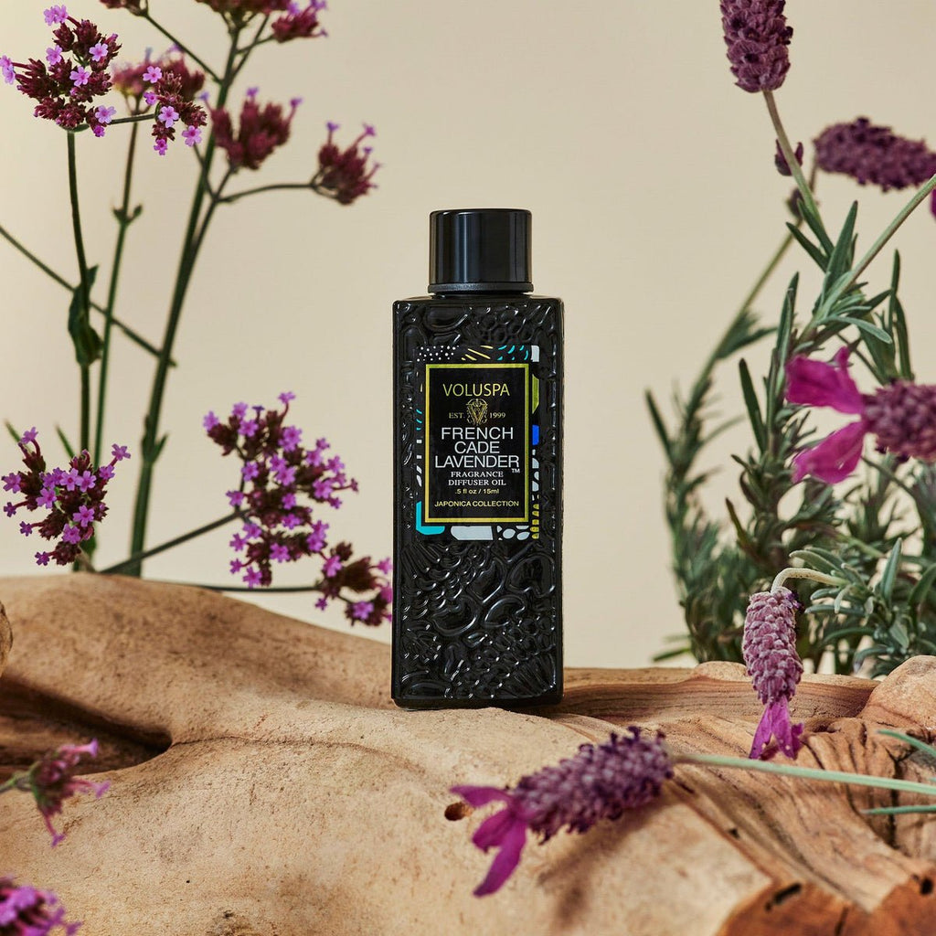 A Voluspa French Cade Lavender Ultrasonic Diffuser Oil bottle rests on French Cade wood, surrounded by lavender sprigs and purple flowers, creating a soothing natural ambiance.