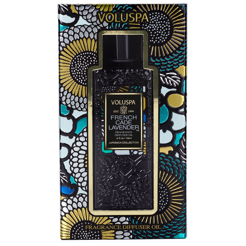 The Voluspa - French Cade Lavender Ultrasonic Diffuser Oil soothes your senses with its aroma. The package features intricate floral designs in black, gold, blue, and green, partially revealing a dark bottle adorned with detailed patterns reminiscent of French Cade Wood.