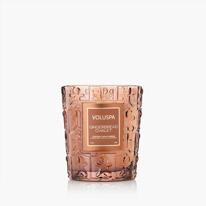 The Voluspa - Gingerbread Chalet Classic 6.5oz Candle features a pink-tinted, textured glass design and coconut wax blend, offering a festive holiday scent from Voluspa, shown against a plain white background.