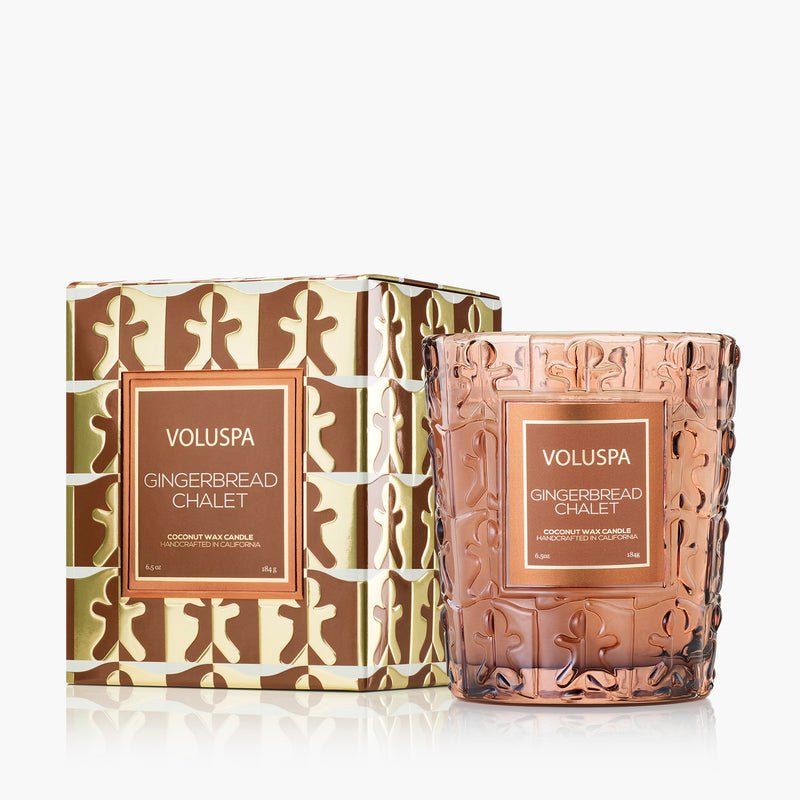 The Voluspa Gingerbread Chalet Classic 6.5oz Candle comes in a textured glass jar with gingerbread figures, paired with a matching gold and brown decorative box featuring a festive gingerbread design.