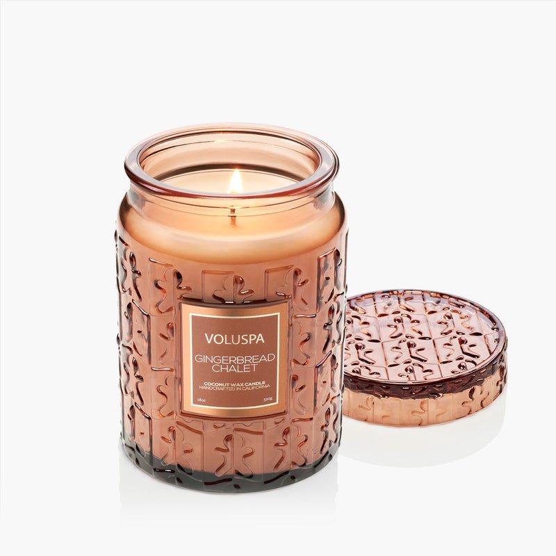 The Voluspa Gingerbread Chalet Large Jar 18oz Candle glows in amber glass with its lid nearby, filling your home with a cozy holiday scent. Crafted from a coconut wax blend by Voluspa, it’s perfect for festive ambiance.