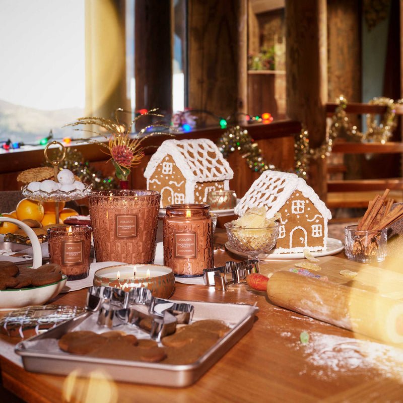 A festive kitchen scene with gingerbread houses, cookies, cookie cutters, spices, baking tools, and holiday lights—accented by the cozy scent of the Voluspa Gingerbread Chalet Large Jar 18oz Candle.