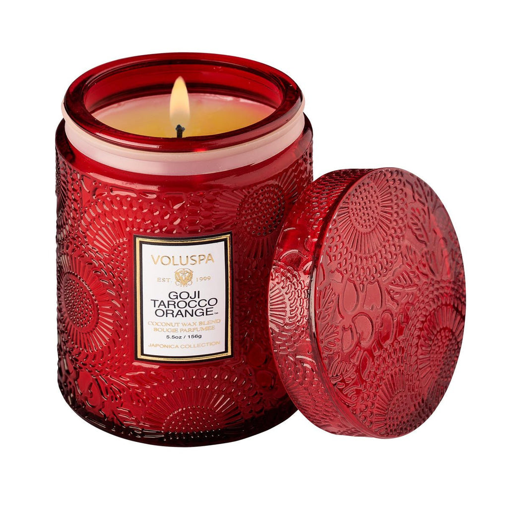 A vibrant red Voluspa Goji Tarocco Orange candle, made from premium coconut wax and housed in an intricately embossed jar, is lit with its lid placed beside, highlighting its decorative design.