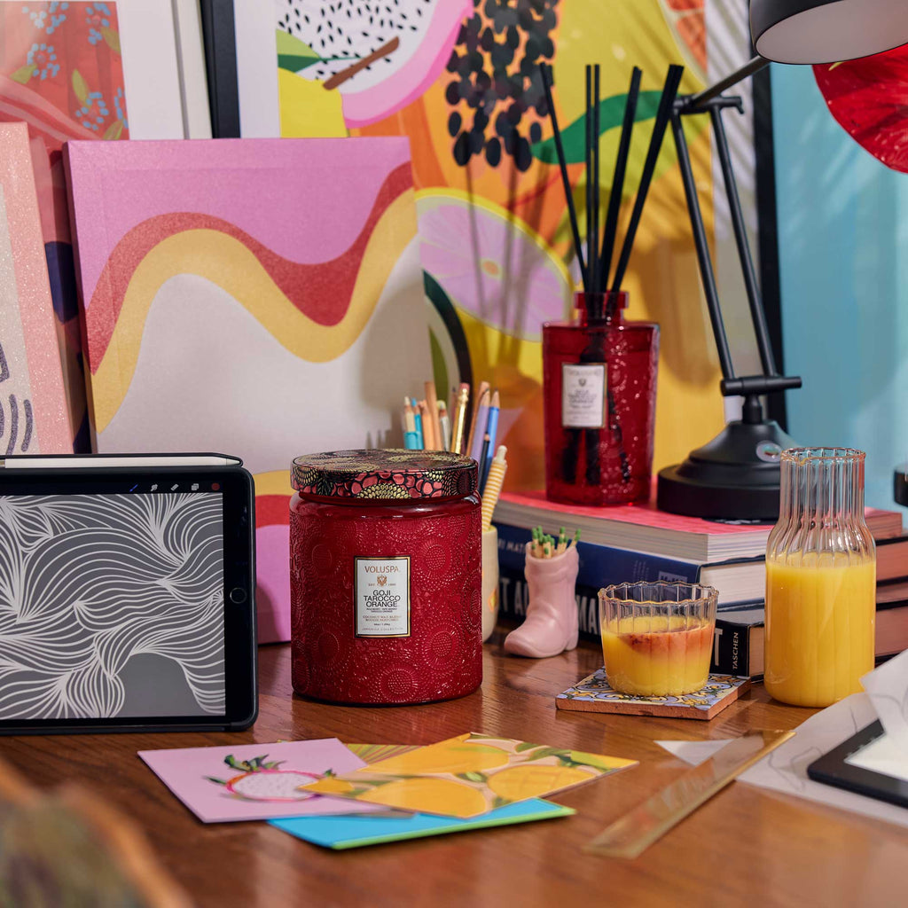 A colorful desk scene includes a Voluspa Goji Torocco Orange Luxe Jar Candle, a tablet with a sketch, juice in glass and bottle, books, and a pink boot-shaped vase with pencils. Bright abstract art decorates the background.