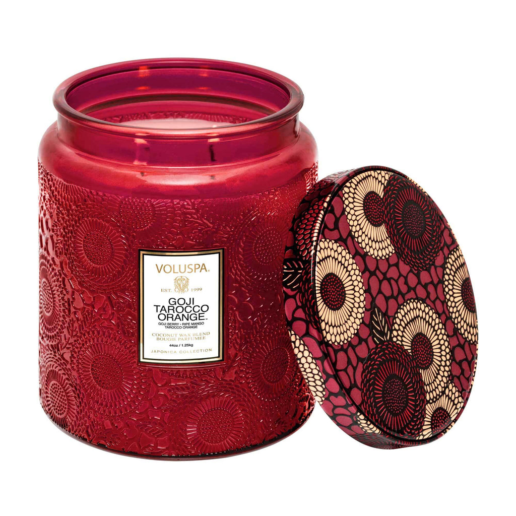 A red glass Voluspa - Goji Torocco Orange Luxe Jar 44oz Candle is displayed with its lid off, showcasing a coconut wax blend. The lid features a black, cream, and gold floral pattern, while the jar has an embossed floral design.