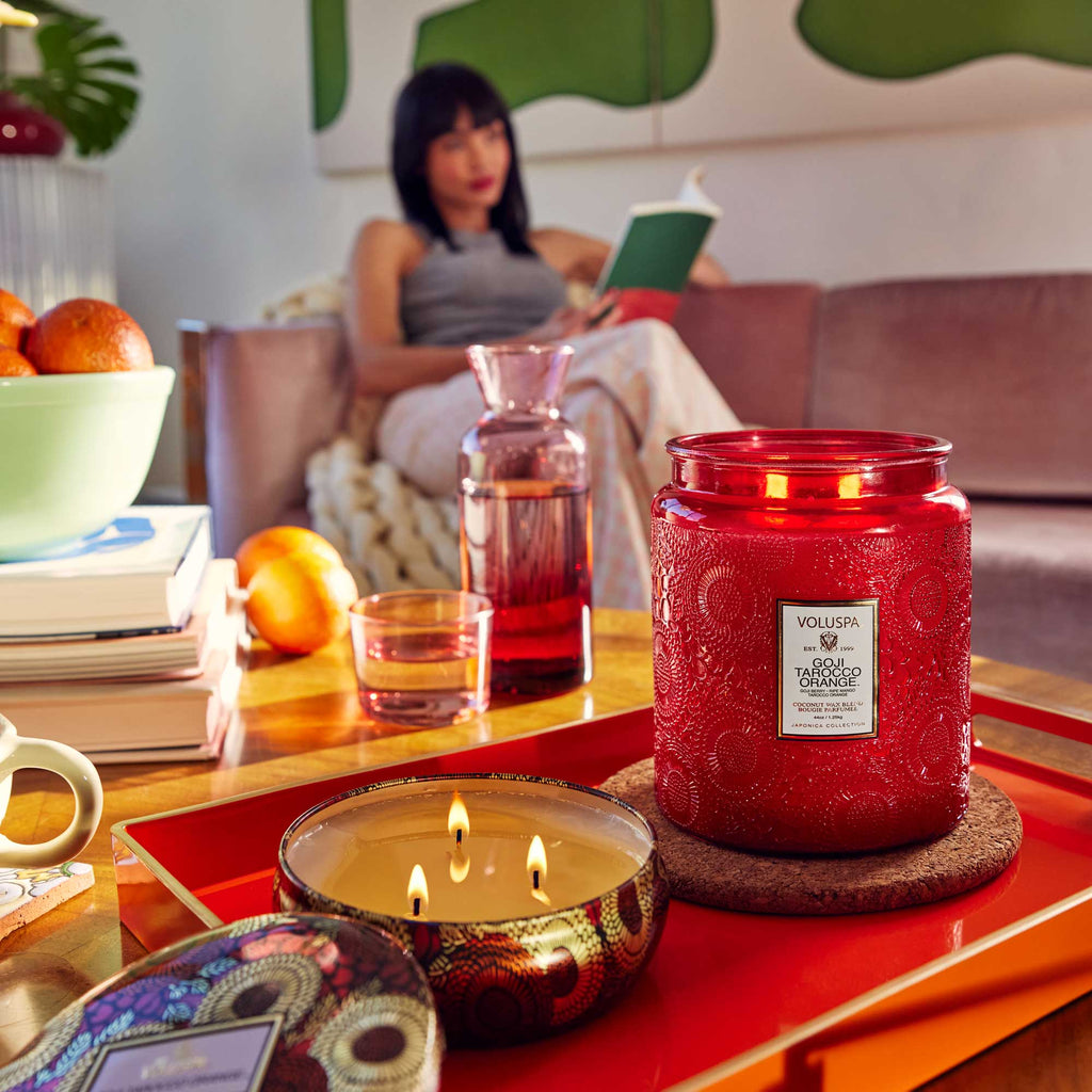 In a vibrant living room, the Voluspa Goji Torocco Orange Luxe Jar 44oz candle sits on a wooden table. A woman reads on the sofa in the background, with fruit and a glass pitcher beside her, creating a warm, cozy ambiance.