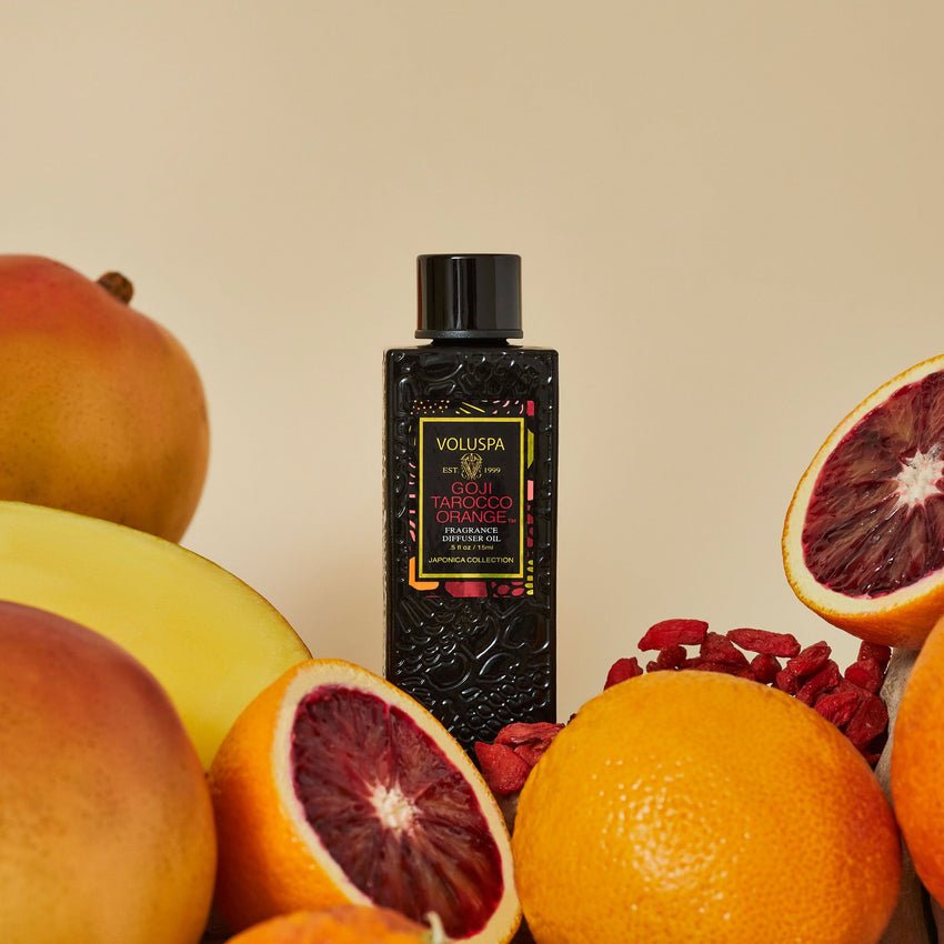 A black Voluspa bottle labeled Goji Torocco Orange Ultrasonic Diffuser Oil and an ultrasonic diffuser sit amid fresh blood oranges, mango, and goji berries on a beige background, accentuating the vibrant fruity theme.