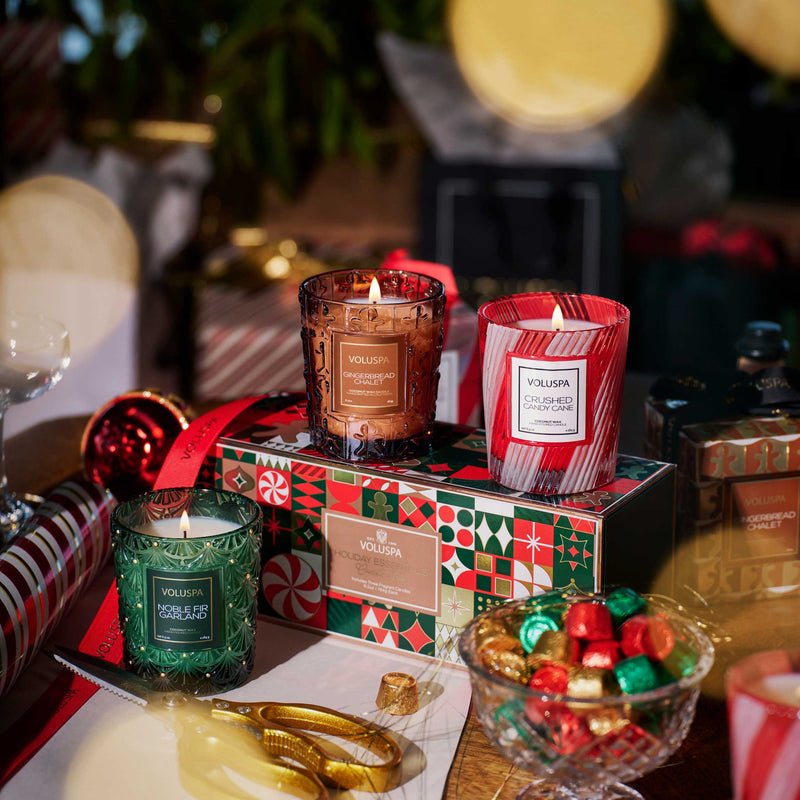 The Voluspa - Holiday Essentials Candle Trio Gift Set features three festive candles in decorative glass jars, beautifully arranged with a patterned box—an ideal holiday gift set to brighten up any seasonal celebration.