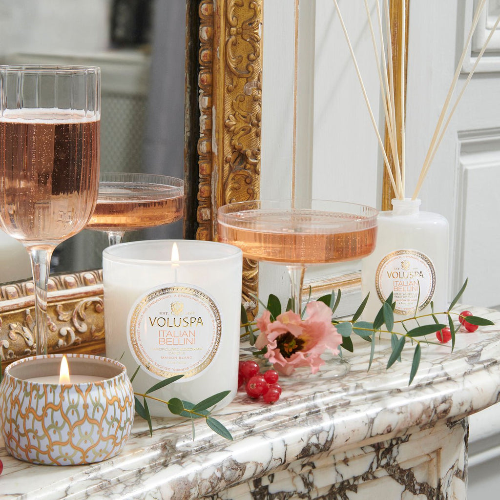 A candle-lit marble scene features elegant Voluspa candles, two glasses of sparkling rosé, and a Voluspa Italian Bellini Reed Diffuser. Decorative flowers and red berries add natural charm, while a mirror reflects the arrangements allure, enhanced by a hint of white peach.