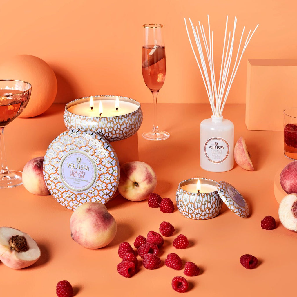 A Voluspa Italian Bellini Reed Diffuser and lit candles sit on an orange surface, surrounded by peaches, raspberries, and champagne glasses, creating a warm and inviting ambiance with the diffusers white peach aroma.